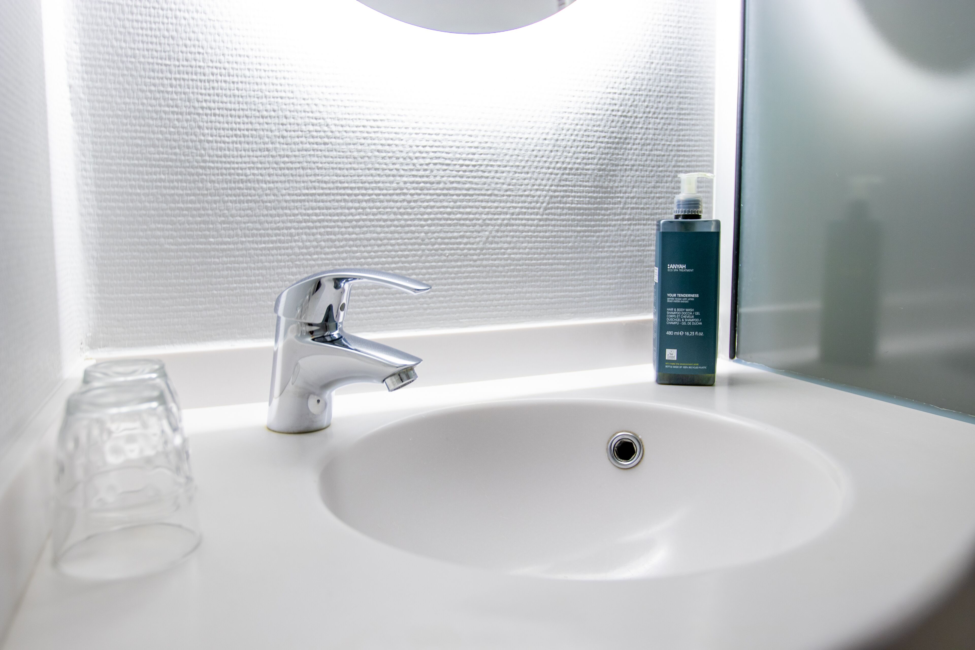 Un lavabo blanc dans une salle de bain, avec un robinet chromé, un verre et une bouteille de shampoing.