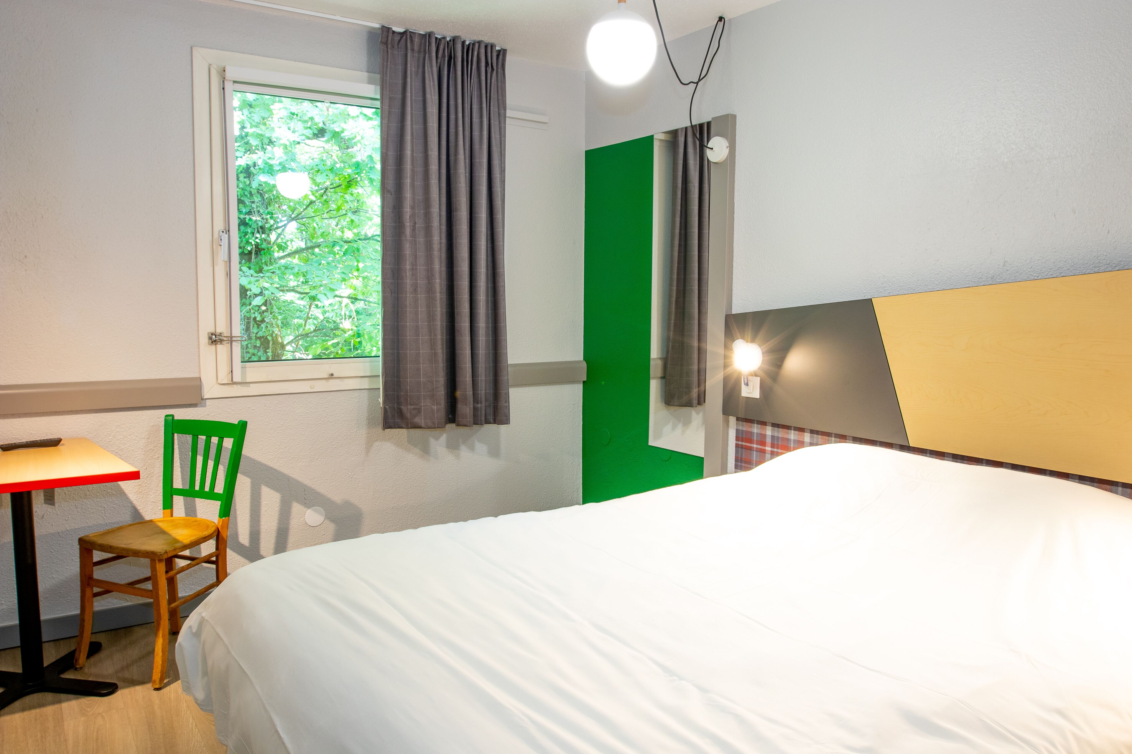 A hotel room with a bed, a chair, a table, a window with curtains, and a green accent wall.
