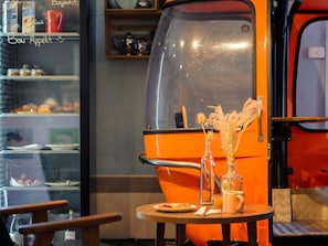 A café with a vintage orange espresso machine, a small table with a plate of food, and a glass vase with dried flowers.