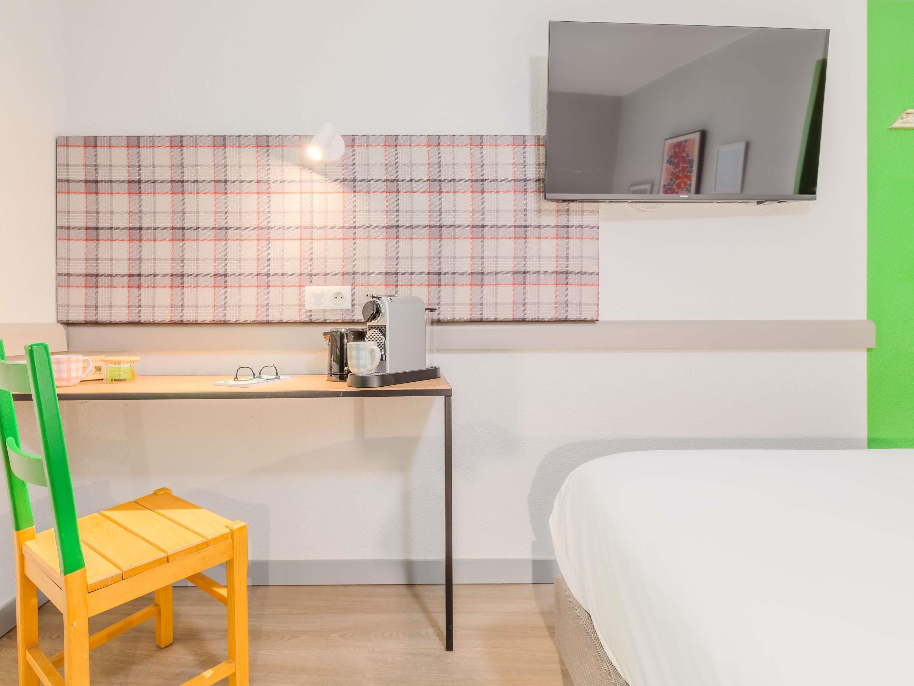Una habitación de hotel con una cama, un escritorio con cafetera, una silla, un televisor y un tapiz de pared a cuadros.