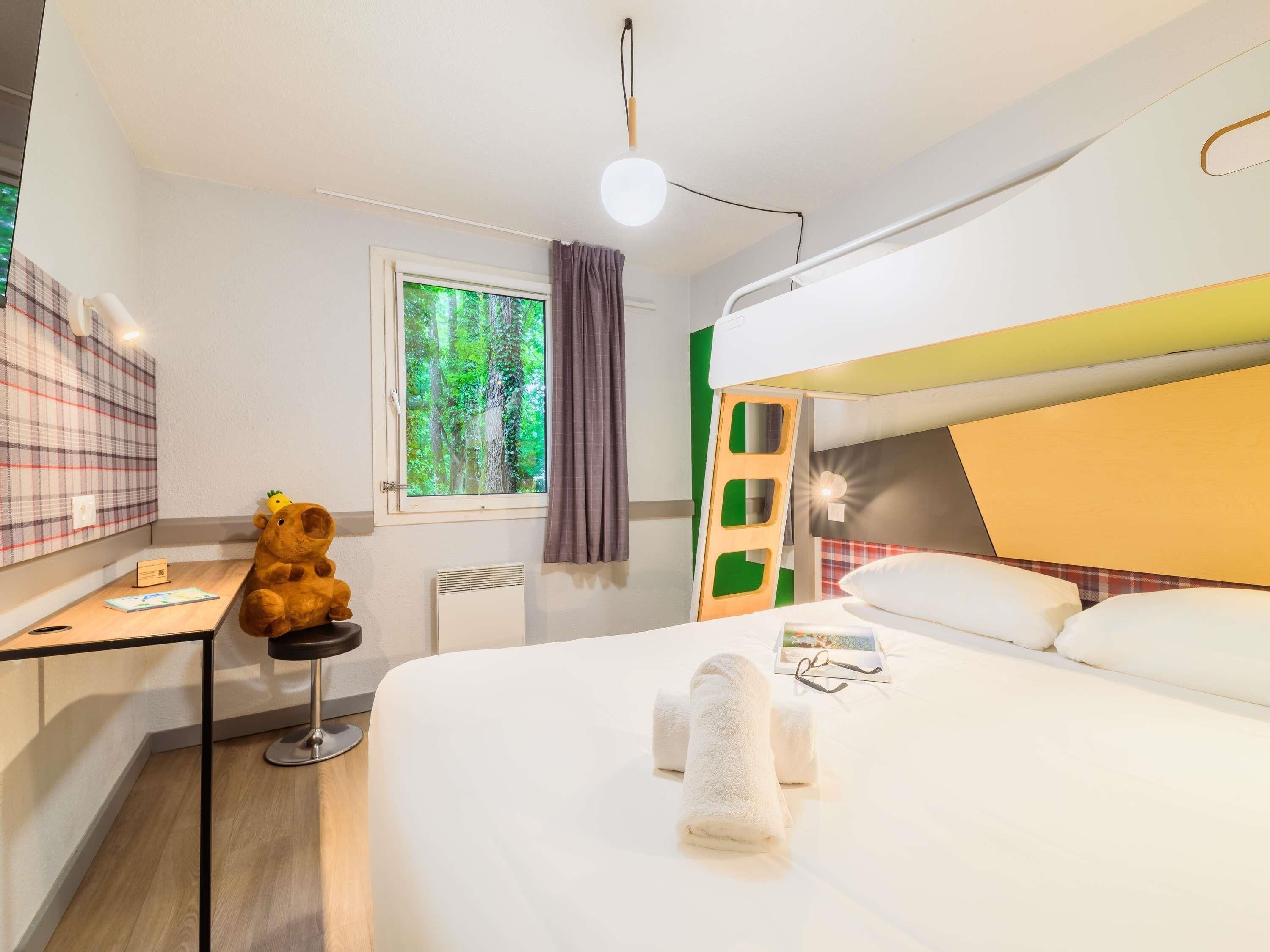 Une chambre d’hôtel avec un lit superposé, un bureau avec une chaise, un ours en peluche, une fenêtre donnant sur des arbres et une table de chevet avec un livre et des lunettes.