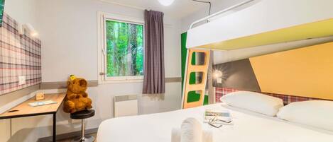 Une chambre d’hôtel avec un lit superposé, un bureau avec une chaise, un ours en peluche, une fenêtre donnant sur des arbres et une table de chevet avec un livre et des lunettes.