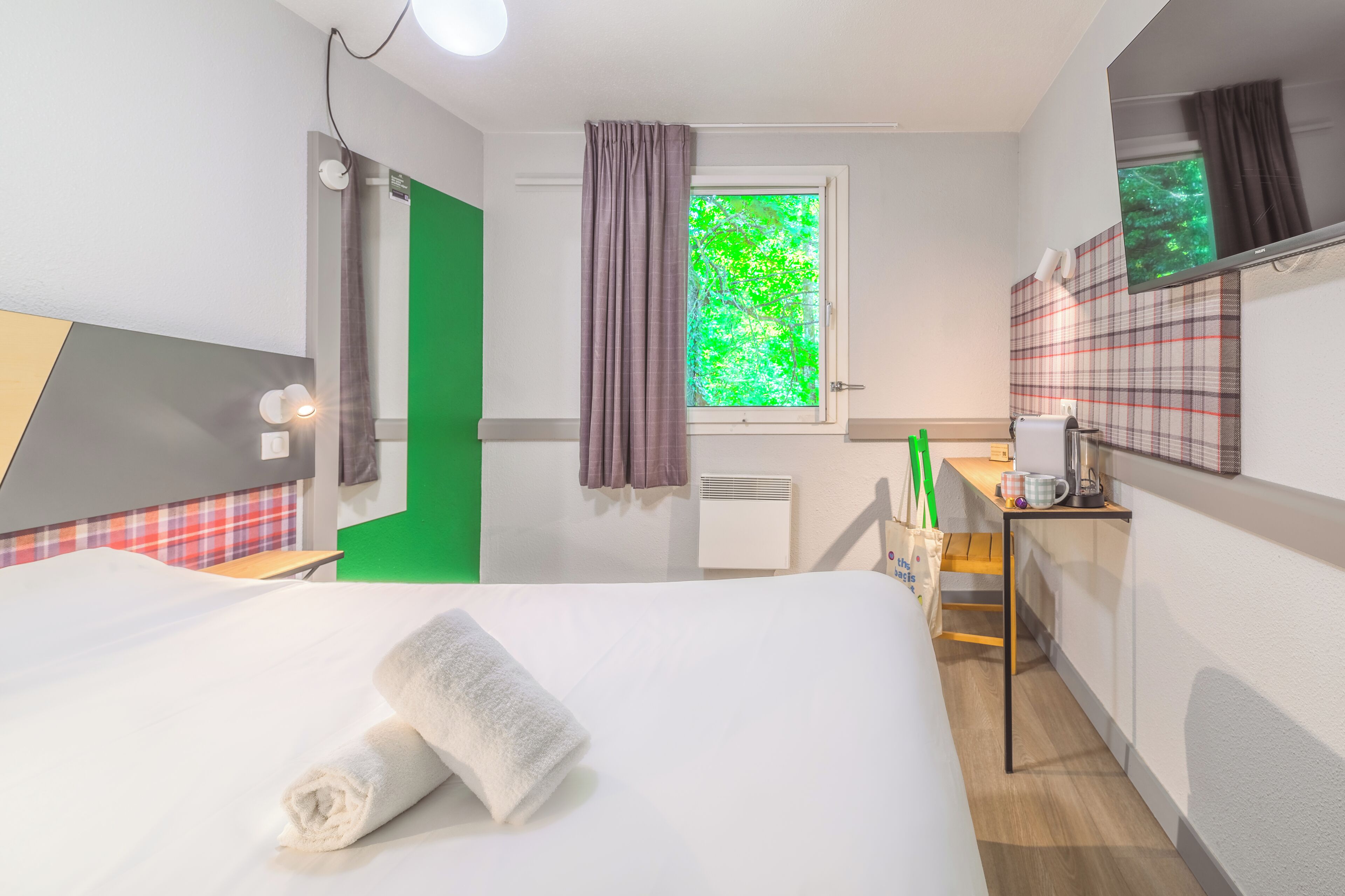 A hotel room with a bed, a small desk, a window with curtains, a wall-mounted heater, and a green accent wall.