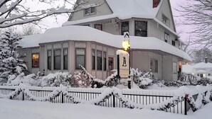 Una casa grande de dos pisos con techo pronunciado, rodeada de árboles cubiertos de nieve y una cerca.