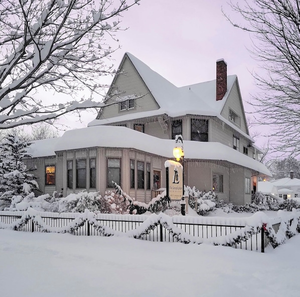 A large, two-story house with a steep roof, surrounded by snow-covered trees and a fence.