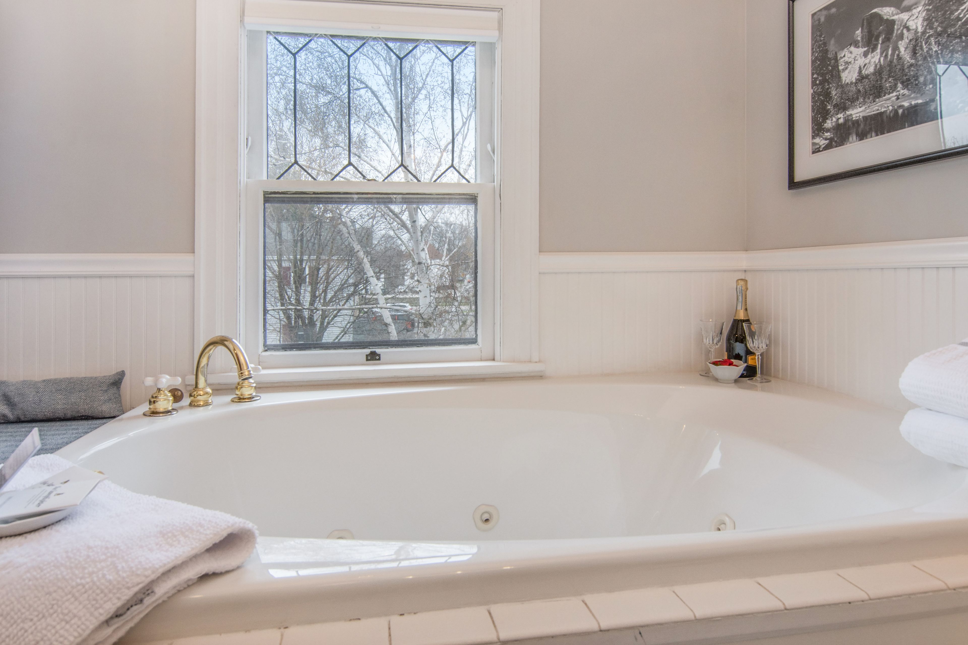 A large white bathtub with a gold faucet, a window with a view of trees, and a framed picture on the wall.