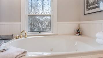 A large white bathtub with a gold faucet, a window with a view of trees, and a framed picture on the wall.