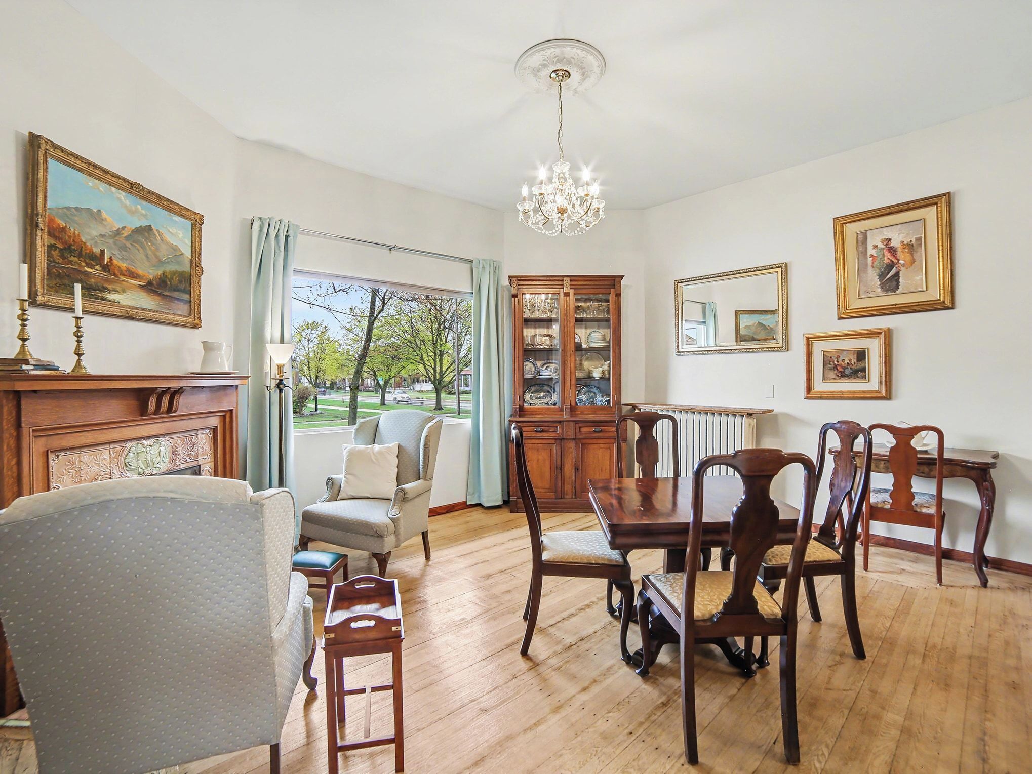 Une pièce avec une cheminée, une table à manger avec des chaises, un lustre et une vue sur une rue bordée d’arbres.