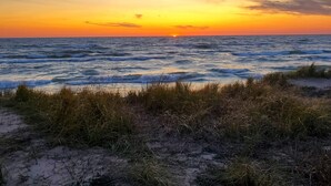 Escena de playa con un atardecer sobre el océano, dunas de arena y vegetación escasa.