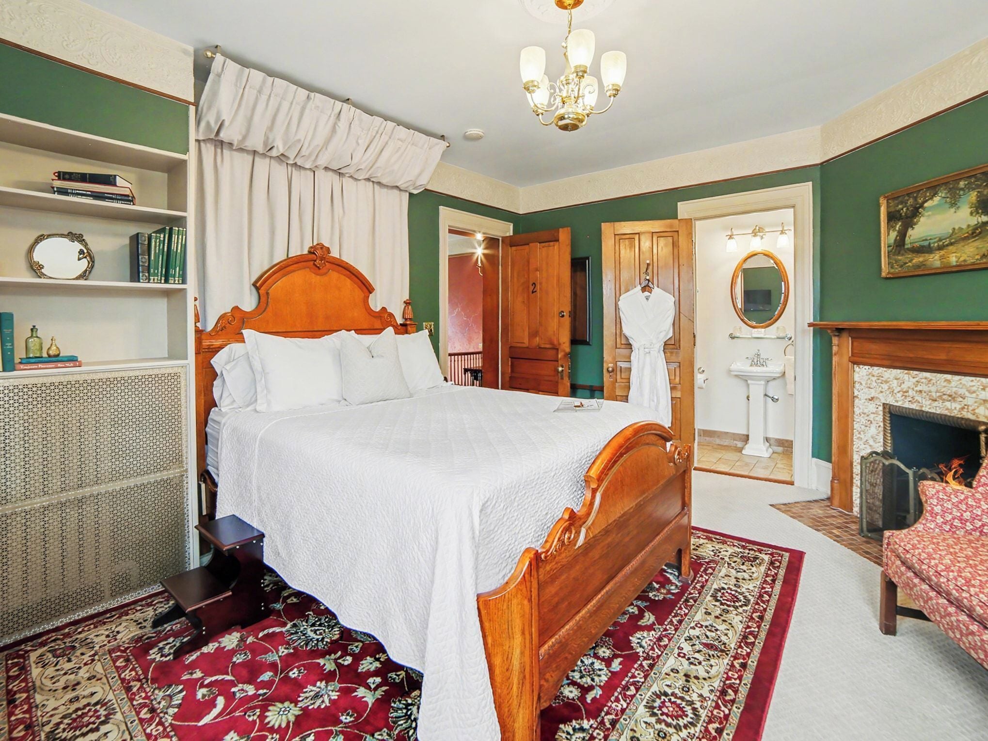 A bedroom with a wooden bed, a red and white patterned rug, a white bedside table, a bookshelf, and a green wall.