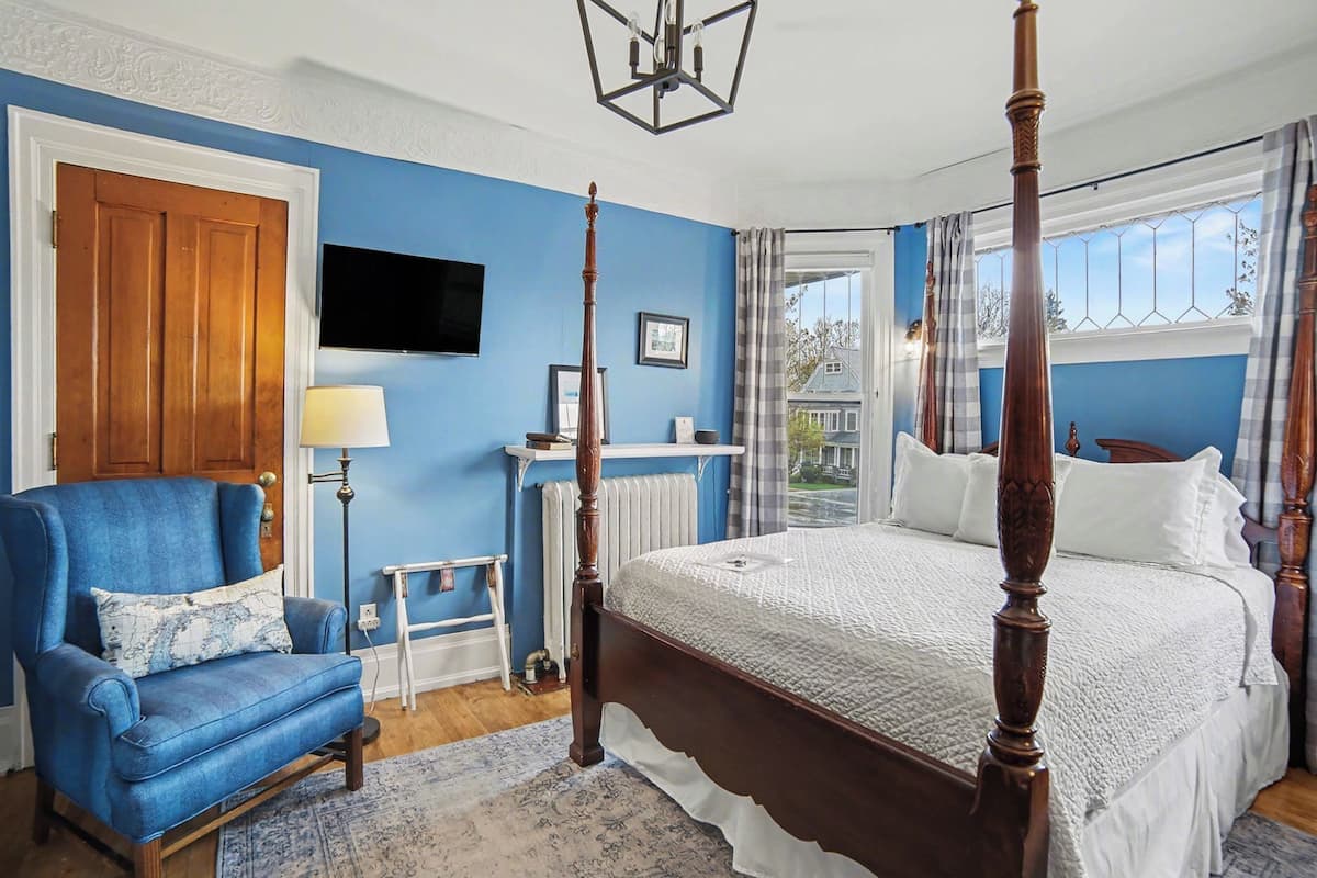 Een slaapkamer met een hemelbed, een blauwe fauteuil, een nachtkastje met een lamp en uitzicht op het omliggende landschap.