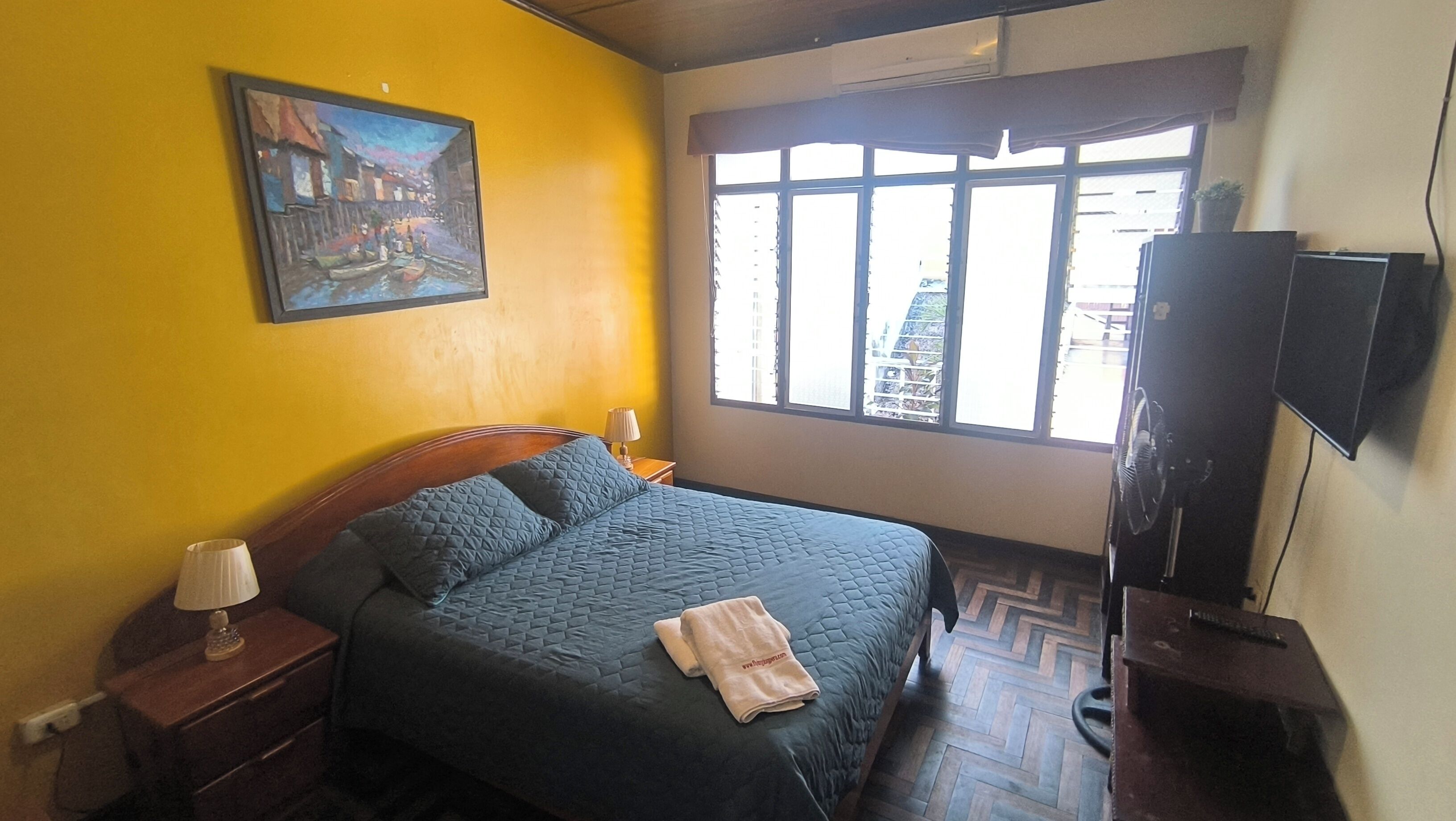 Quarto com cama, mesa de cabeceira, luminária, quadro, janela e televisão.