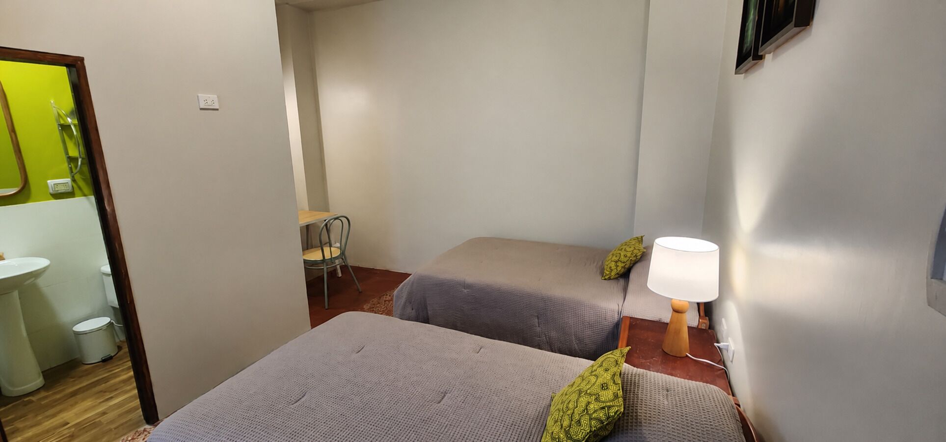 A small, compact room with two single beds, a bedside table with a lamp, a chair, and a small table with a chair.