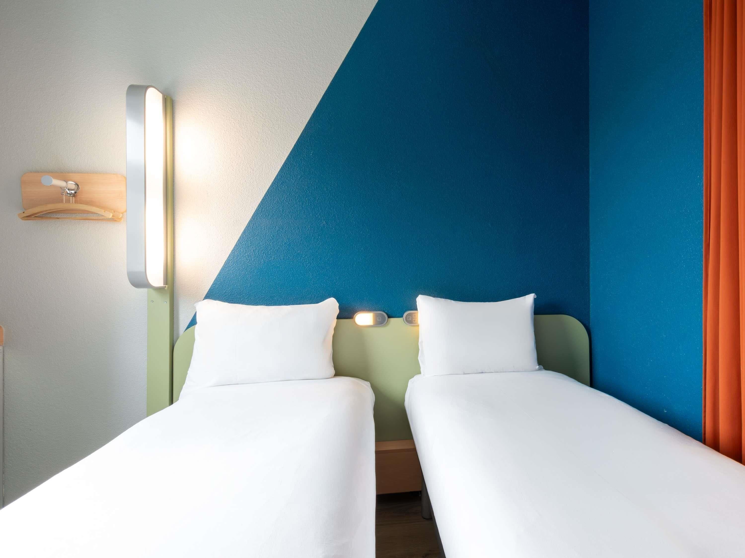 Habitación de hotel con dos camas individuales, una mesita de noche, una lámpara de pared y un perchero de madera.