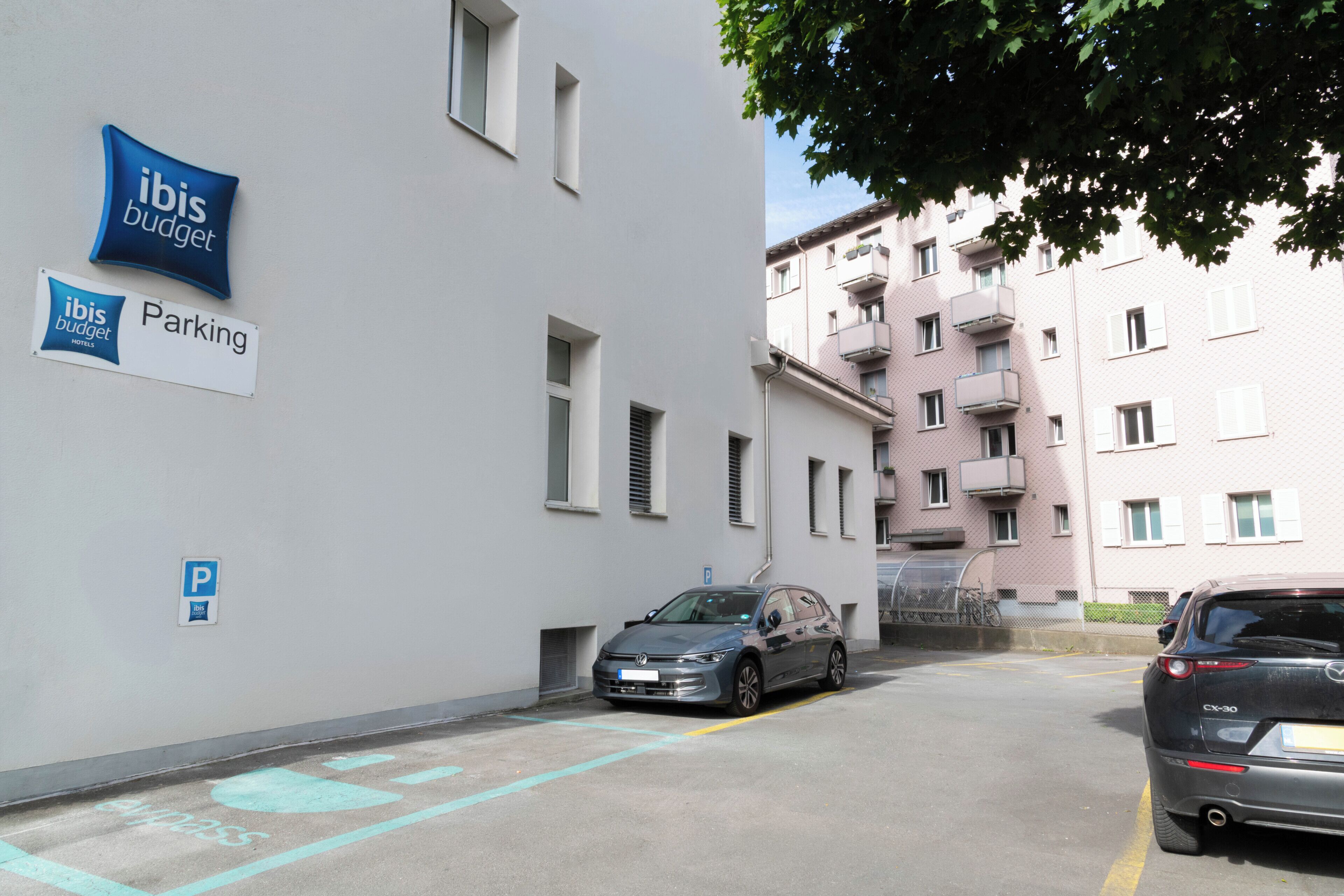 A parking area with a sign for ibis budget hotels, a parked car, and a building with multiple balconies.