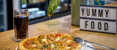 A pizza on a plate with a fork and knife, a glass of dark beverage, and a sign that reads 'YUMMY FOOD' on a table.