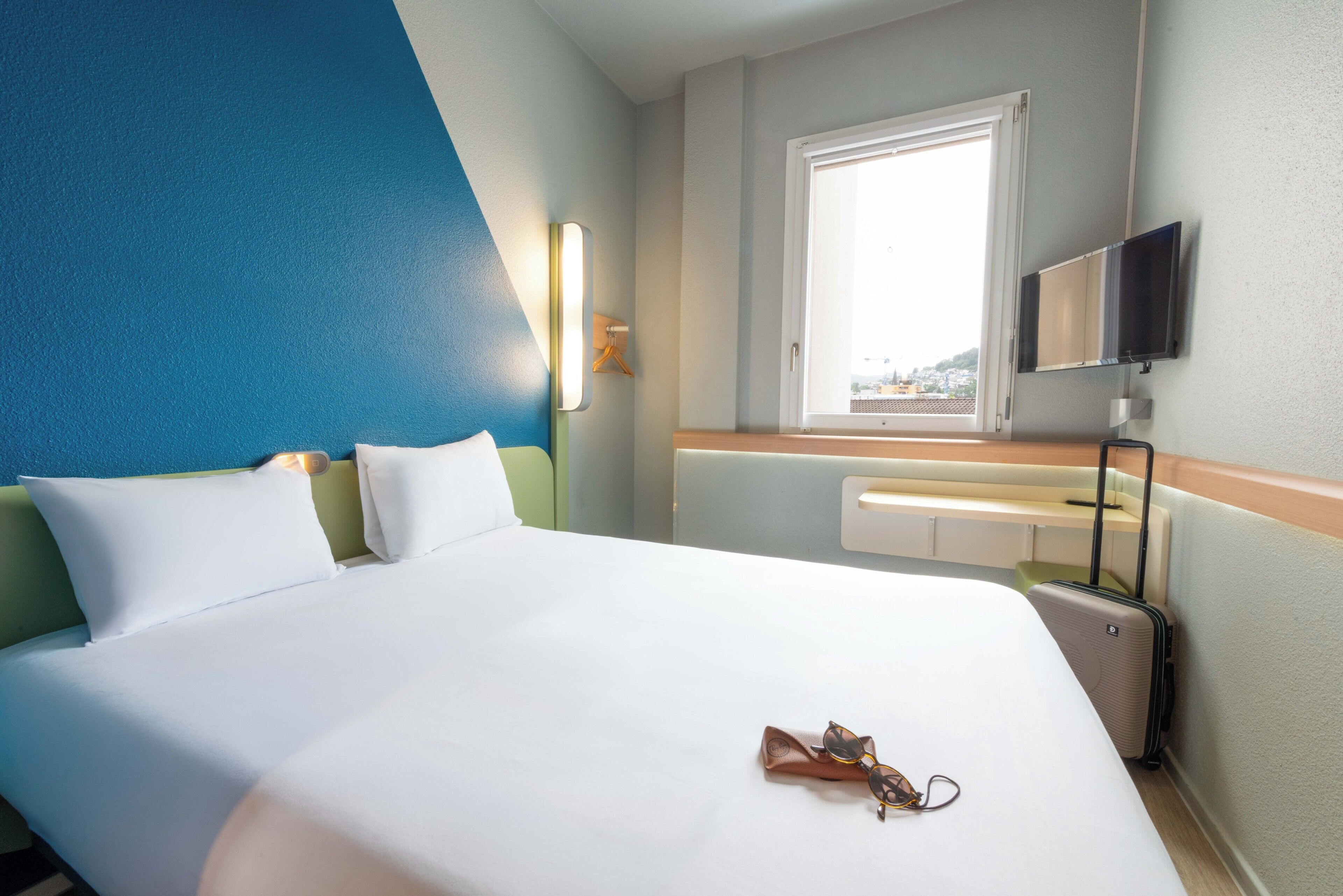A hotel room with a large bed, a television, a window, and a suitcase.