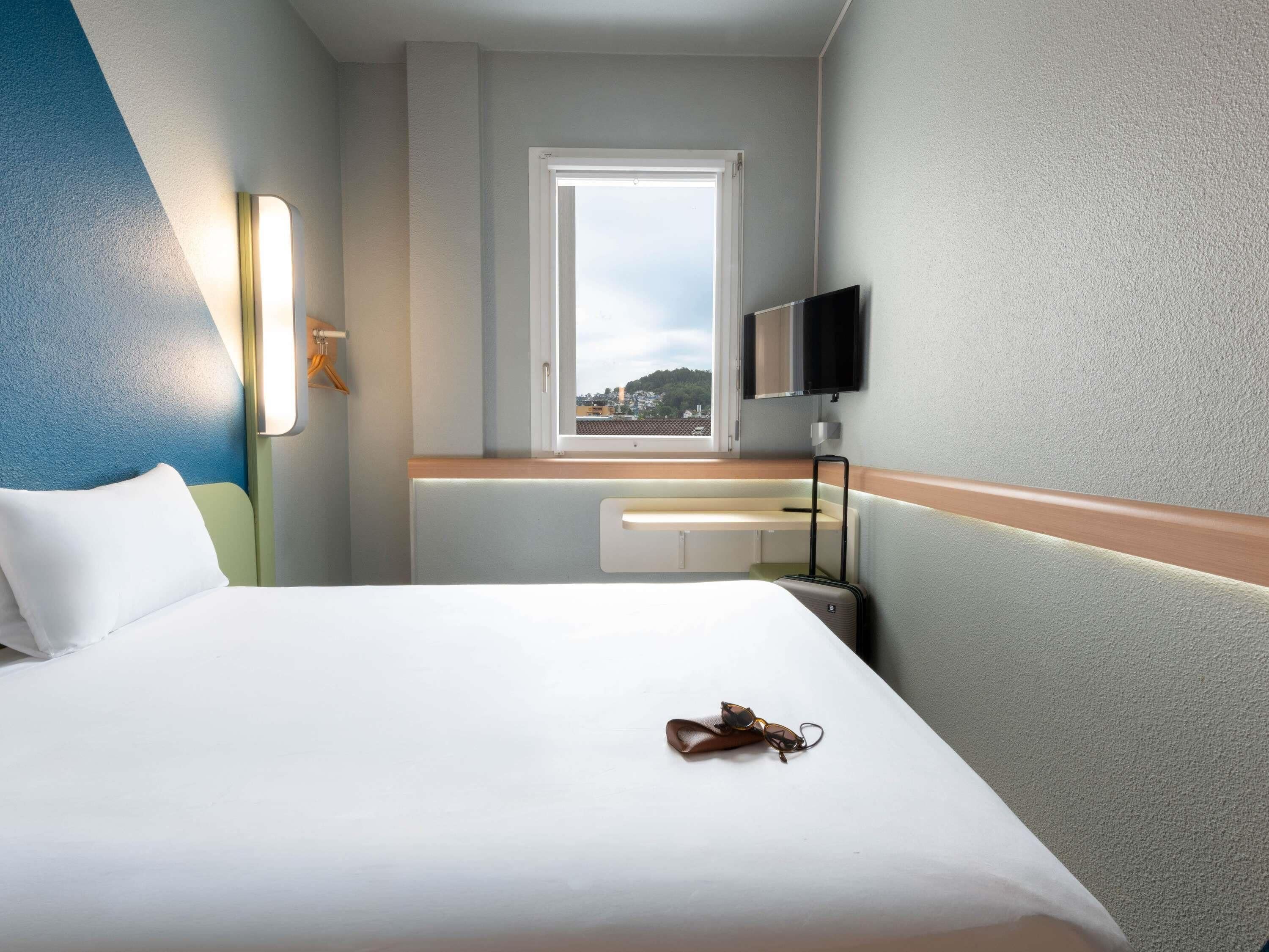 A hotel room with a bed, a window, a television, and a suitcase.