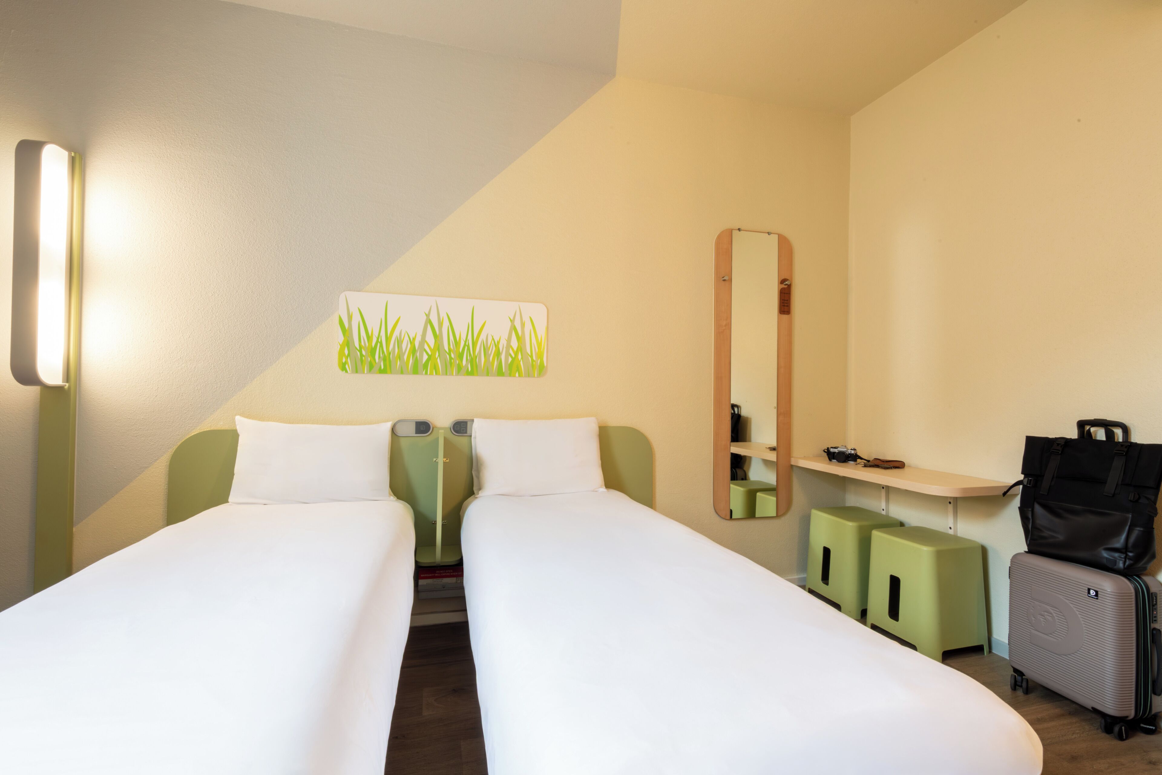 Ein Hotelzimmer mit zwei Einzelbetten, einem Schreibtisch mit Stuhl, einem Spiegel und einem Koffer.