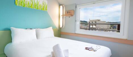 Habitación de hotel con cama, ventana con vistas a edificios y un cuadro de pasto en la pared.