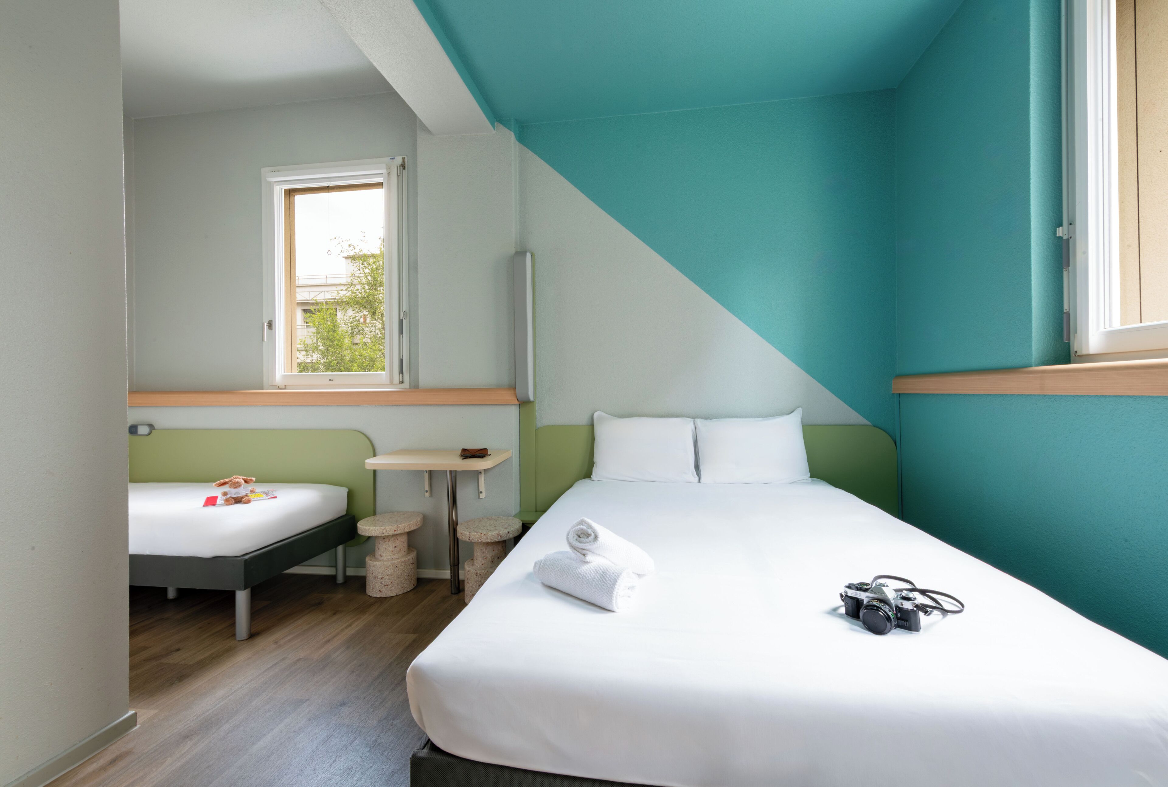Ein Hotelzimmer mit Bett, einem kleinen Tisch, einem Stuhl, einem Fenster und Blick ins Freie.