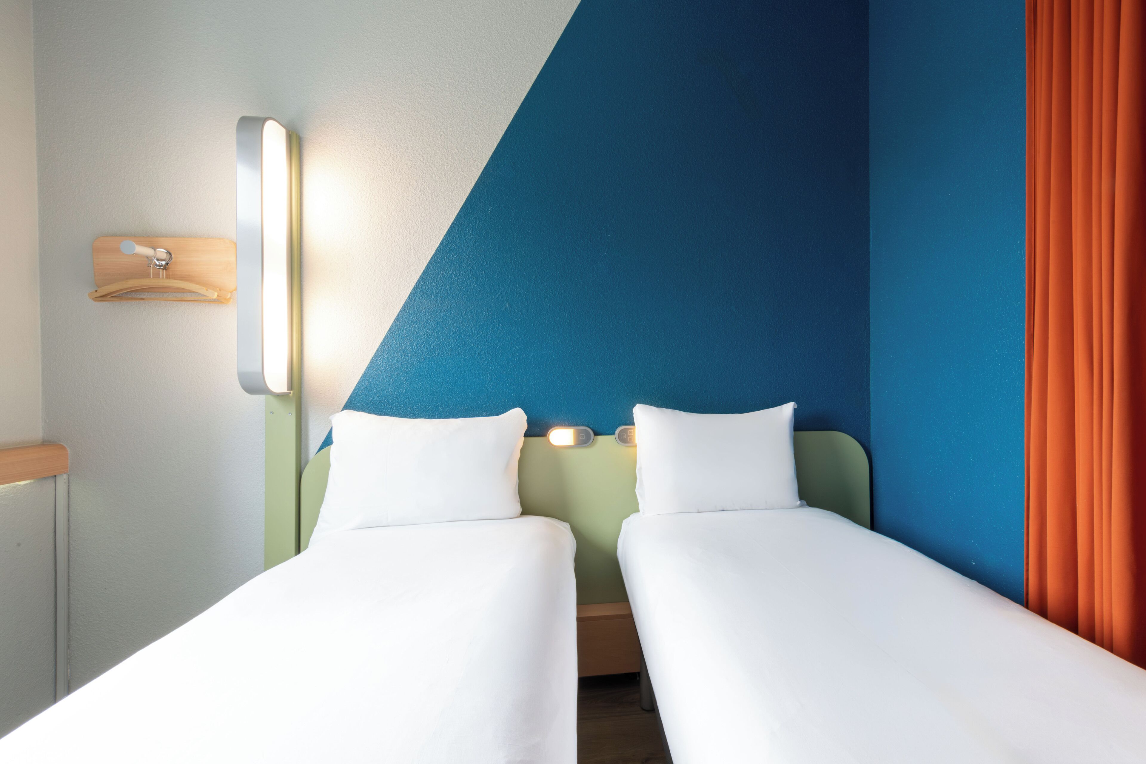 Una camera d'albergo con due letti singoli, un comodino, una lampada a parete e una tenda.