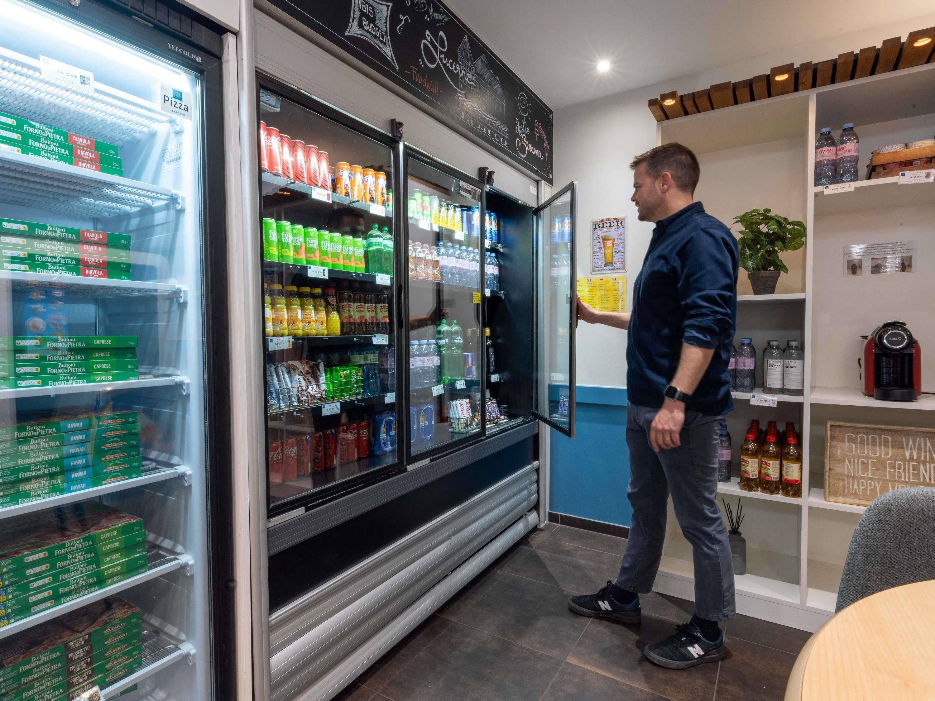 Un homme vêtu d’une chemise bleue et d’un jean se tient devant un réfrigérateur rempli de diverses boissons.