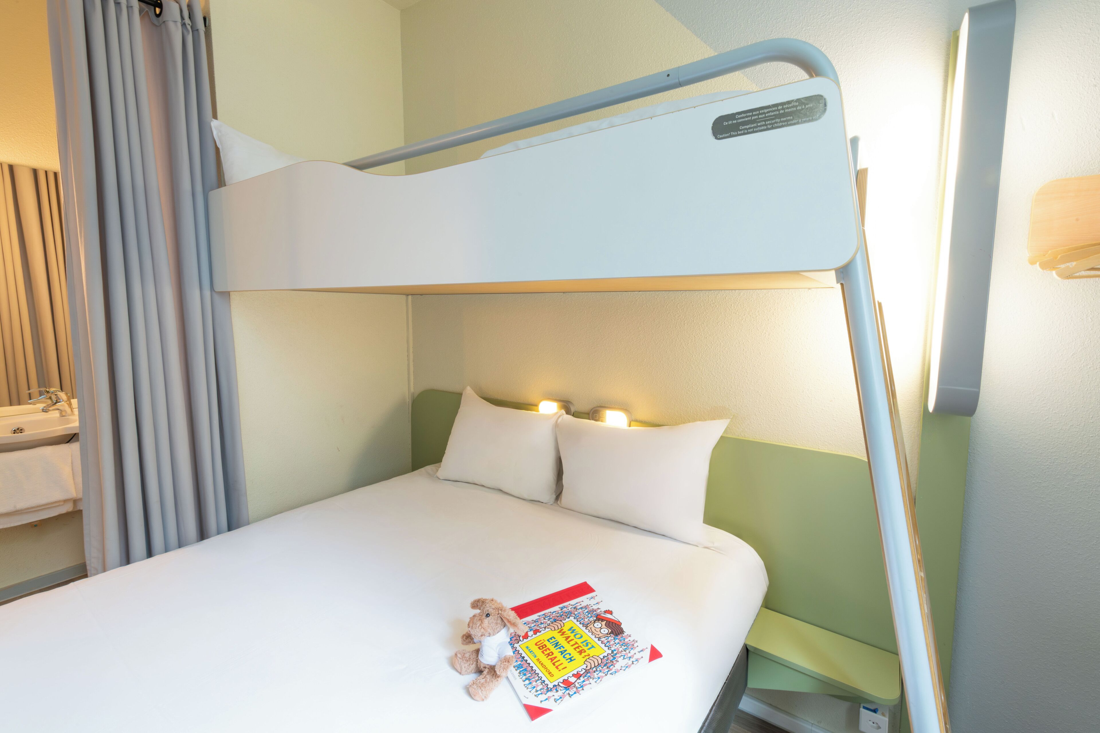 Una habitación de hotel con una cama litera, una mesita de noche con lámpara y un peluche en la cama.