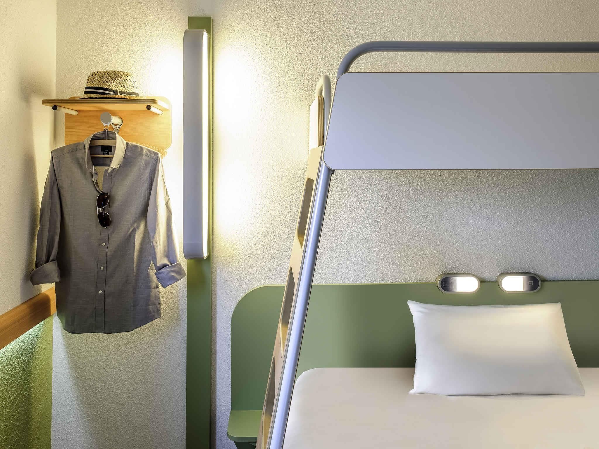 Ein Etagenbett mit grünem Kopfteil und weißem Kopfkissen, ein Kleiderhaken mit grauem Hemd und Sonnenbrille sowie eine wandmontierte Leuchte.
