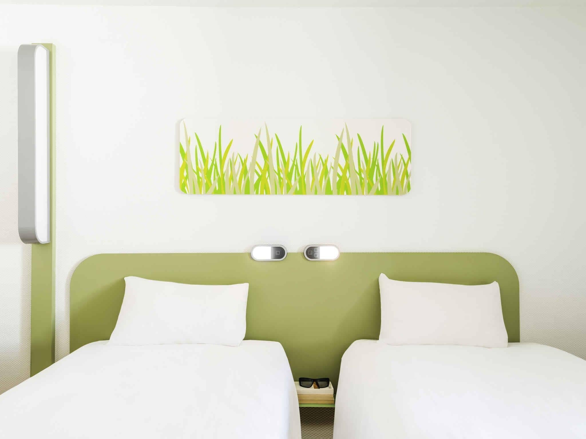 Habitación de hotel con cabecera verde, dos camas blancas, luz adosada a la pared y un cuadro con motivos de hierba.