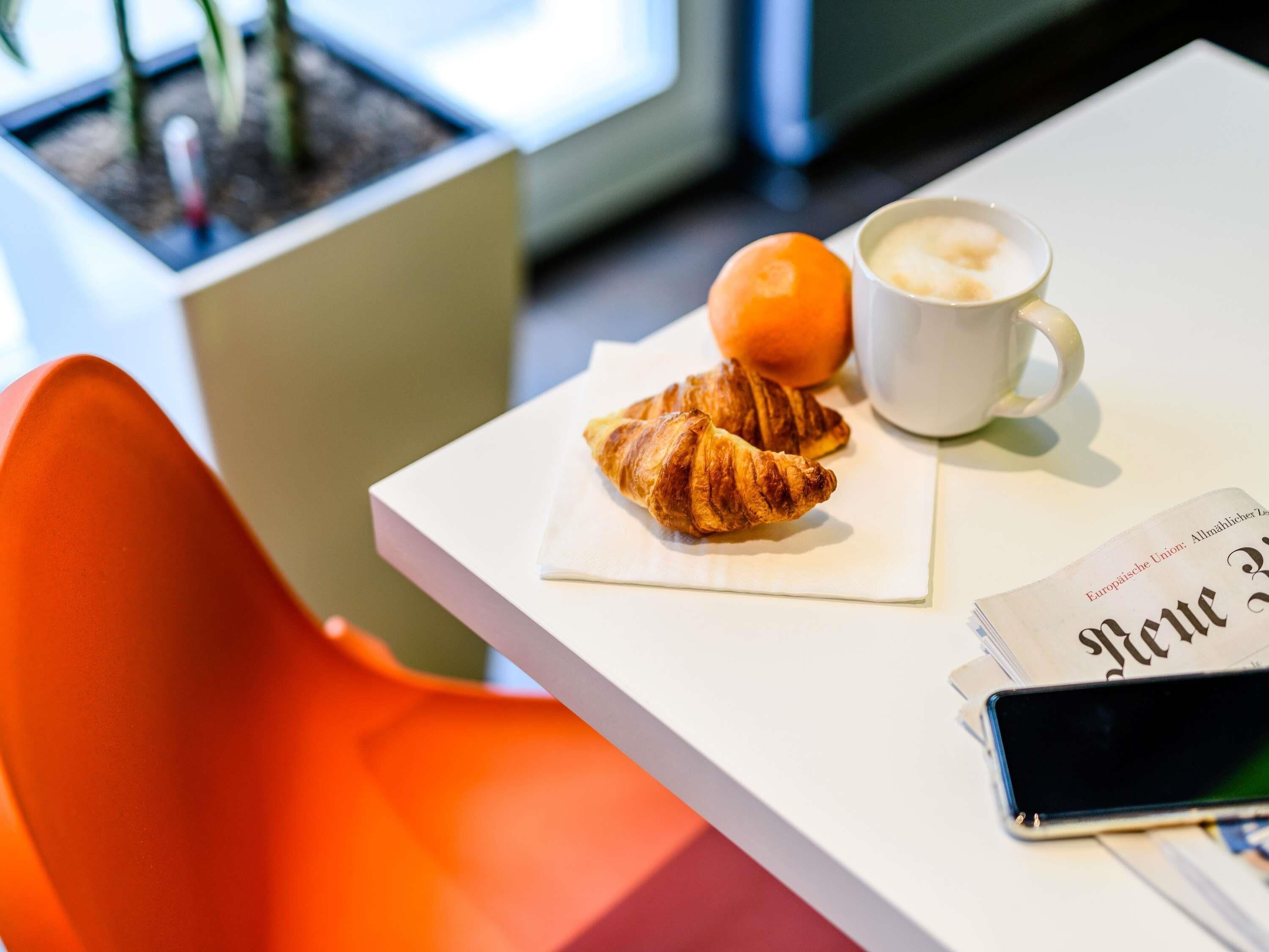 Una mesa con un croissant, una naranja y una taza de café, junto con un periódico y un teléfono inteligente.