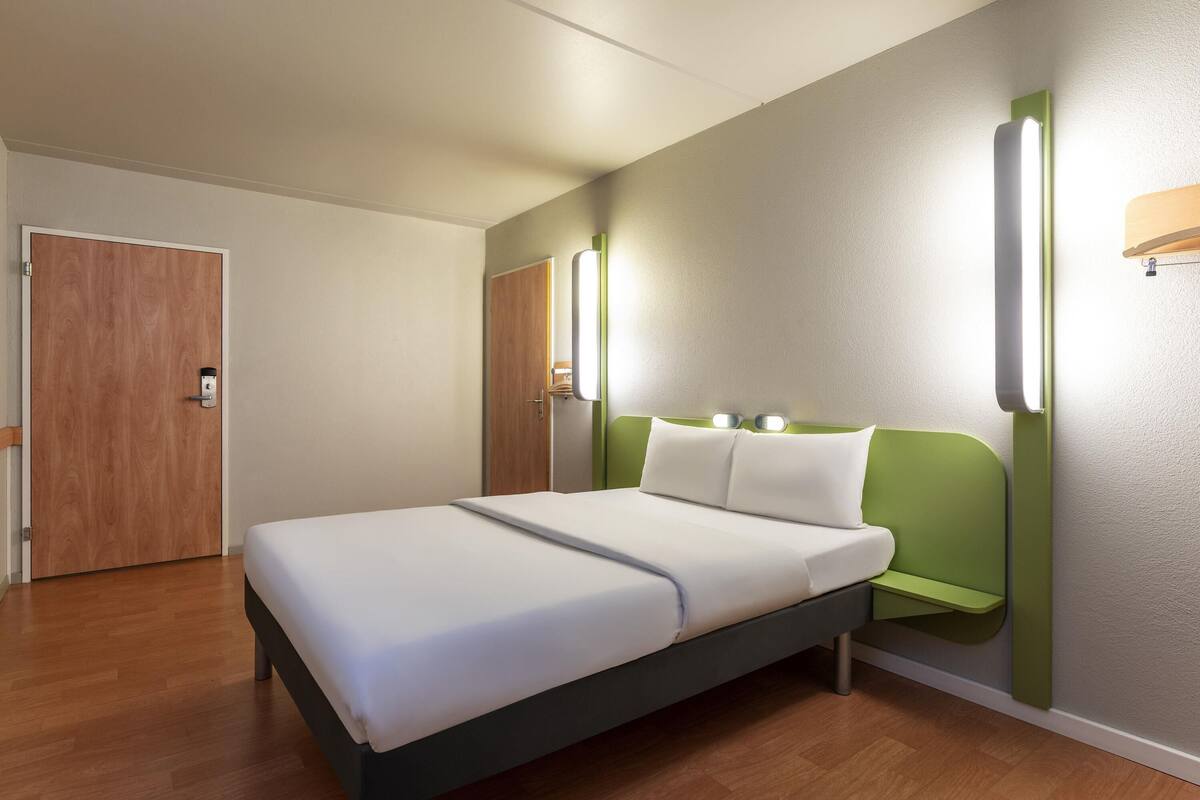 A hotel room with a bed, a door, and a wall-mounted light fixture.