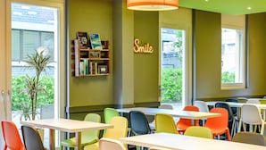 Un área de comedor con sillas y mesas de colores, una estantería con libros, un letrero que dice 'Smile' y vista a zonas verdes al exterior.