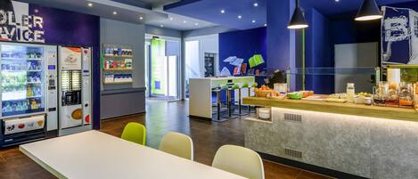 Un café moderno con zona de estar, mostrador con comida y bebidas, y máquinas expendedoras.