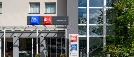 A modern hotel entrance with glass doors, a reception desk, and signage for "ibis budget hotels".