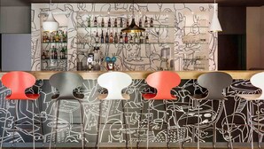 Zona de bar con un mural en la pared, taburetes y una variedad de botellas y vasos en estantes.