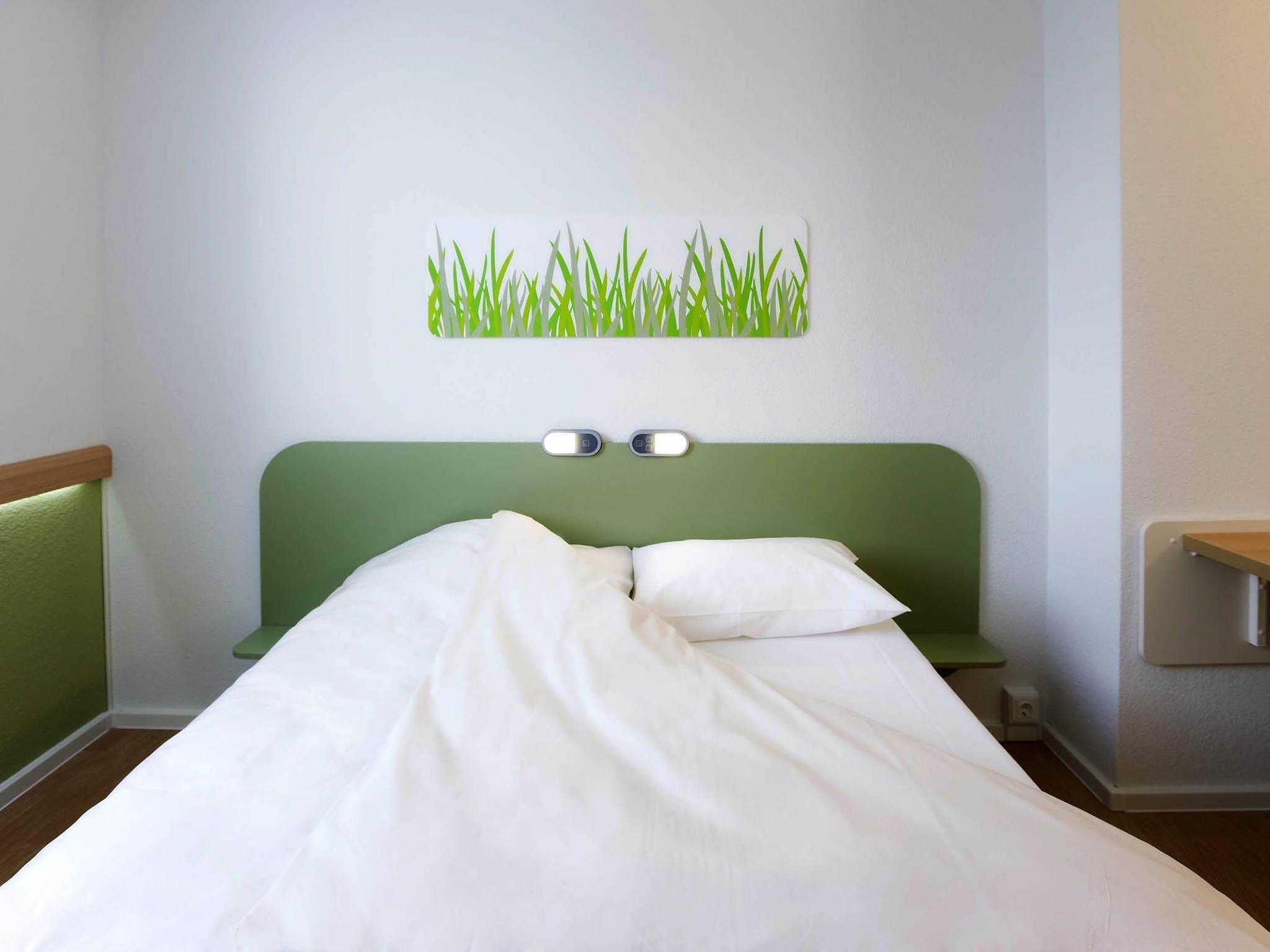 A hotel room with a green headboard, white bedding, and a wall-mounted light fixture.