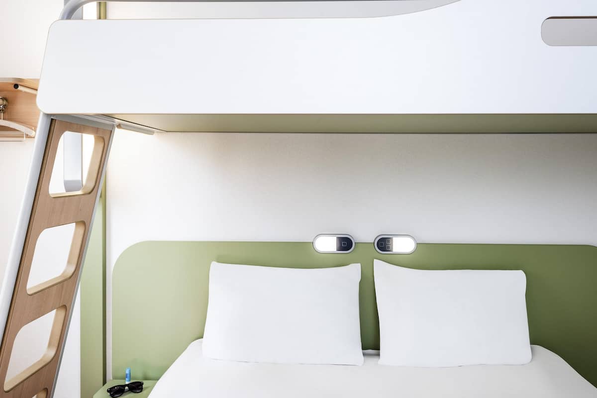 A bunk bed with a green headboard, white bedding, and a small bedside table with a bottle and sunglasses.