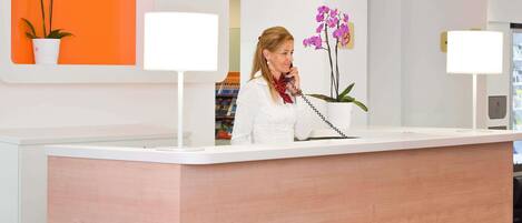 A reception desk with a 'RECEPTION' sign, a potted orchid, and a white wall with a shelf.