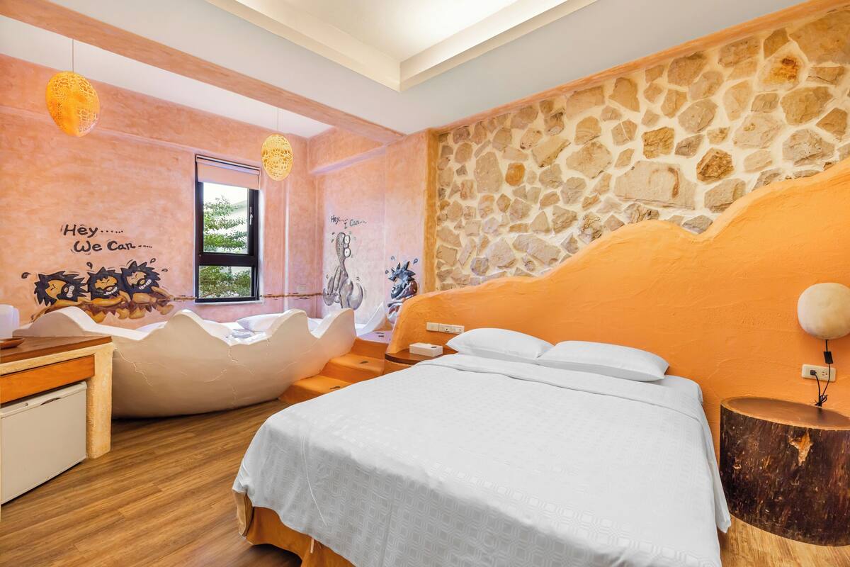Ein modernes Schlafzimmer mit Steinwand, einem großen Bett, einer besonderen Sitzecke und Blick ins Grüne.