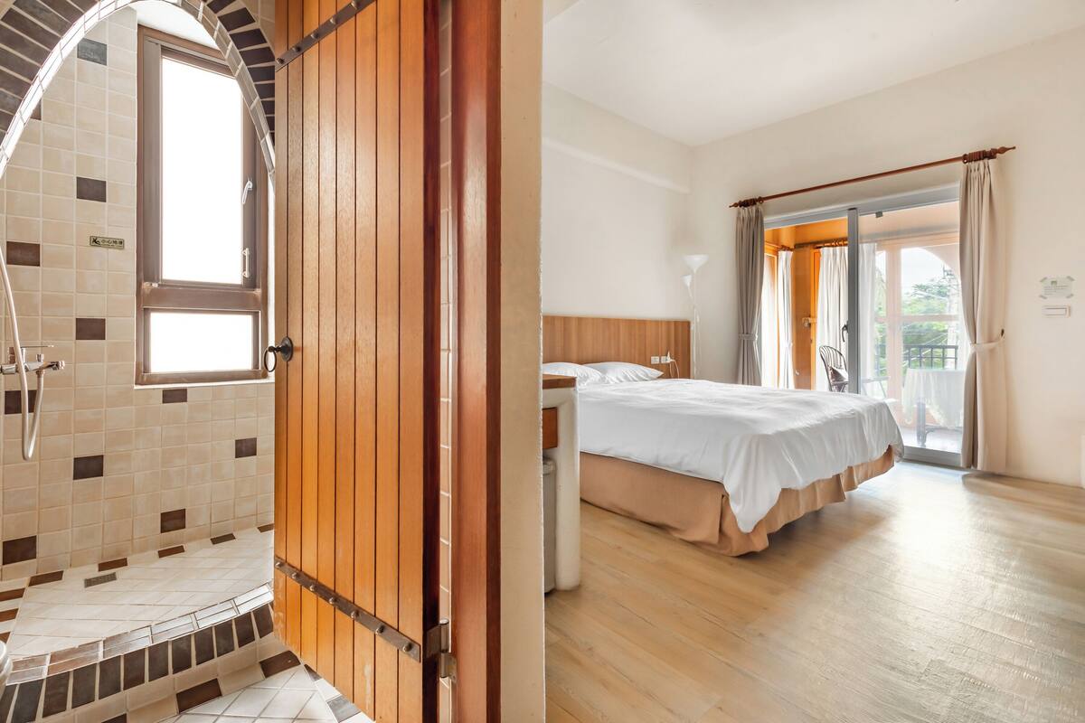 Ein Badezimmer mit Dusche, Toilette und Waschbecken. Eine Holztür mit Fenster. Der Boden und die Wände sind mit Fliesen ausgelegt.