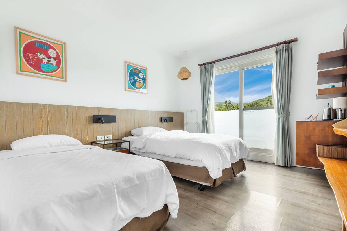 Ein Hotelzimmer mit zwei Betten, einem hölzernen Kopfteil, einem großen Fenster mit Vorhängen und Blick ins Freie.