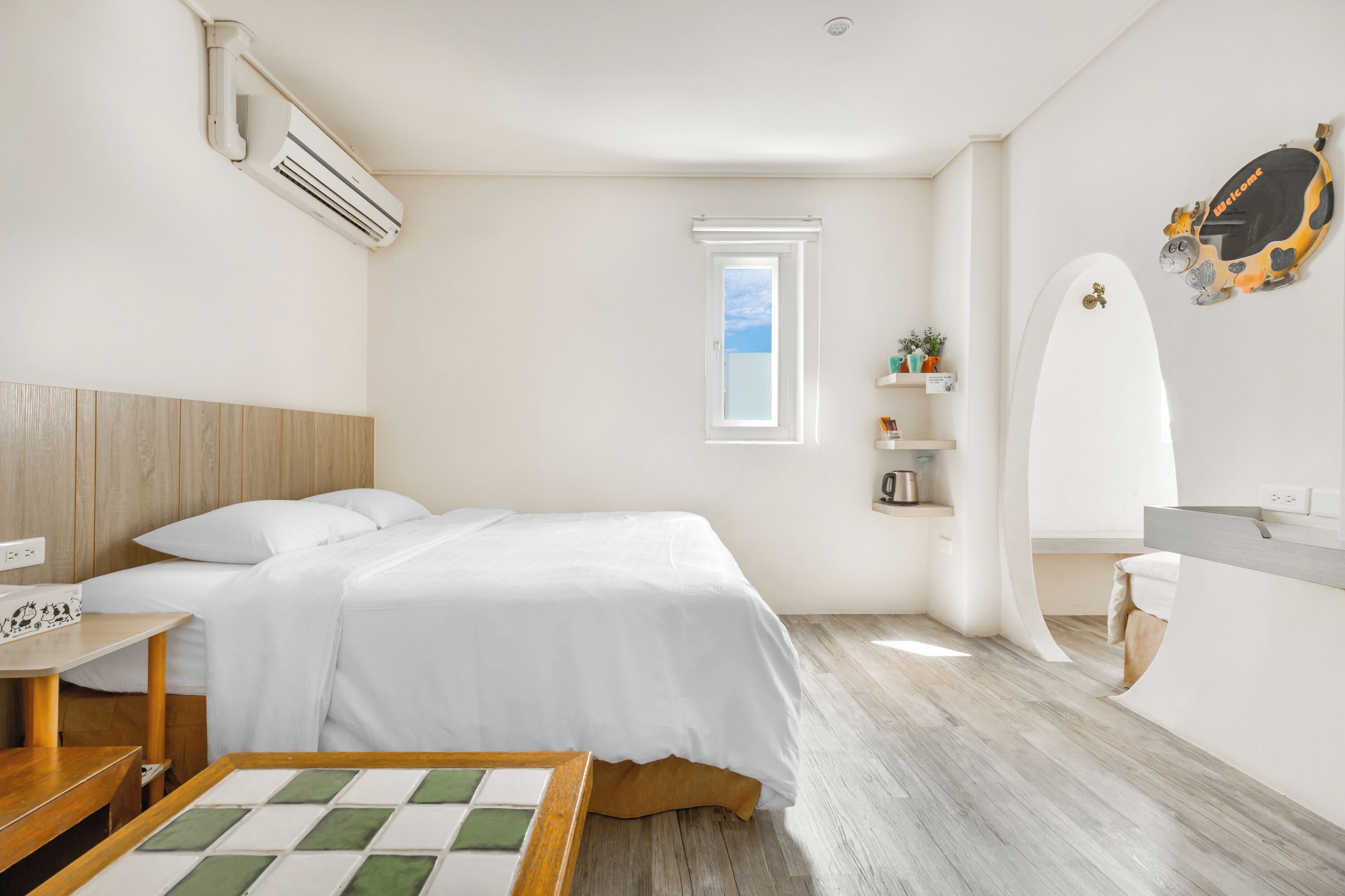 Un dormitorio moderno con cama, mesita de noche y un pequeño escritorio con silla. Se ve una ventana que proporciona luz natural, un aire acondicionado de pared y un adorno en la pared.