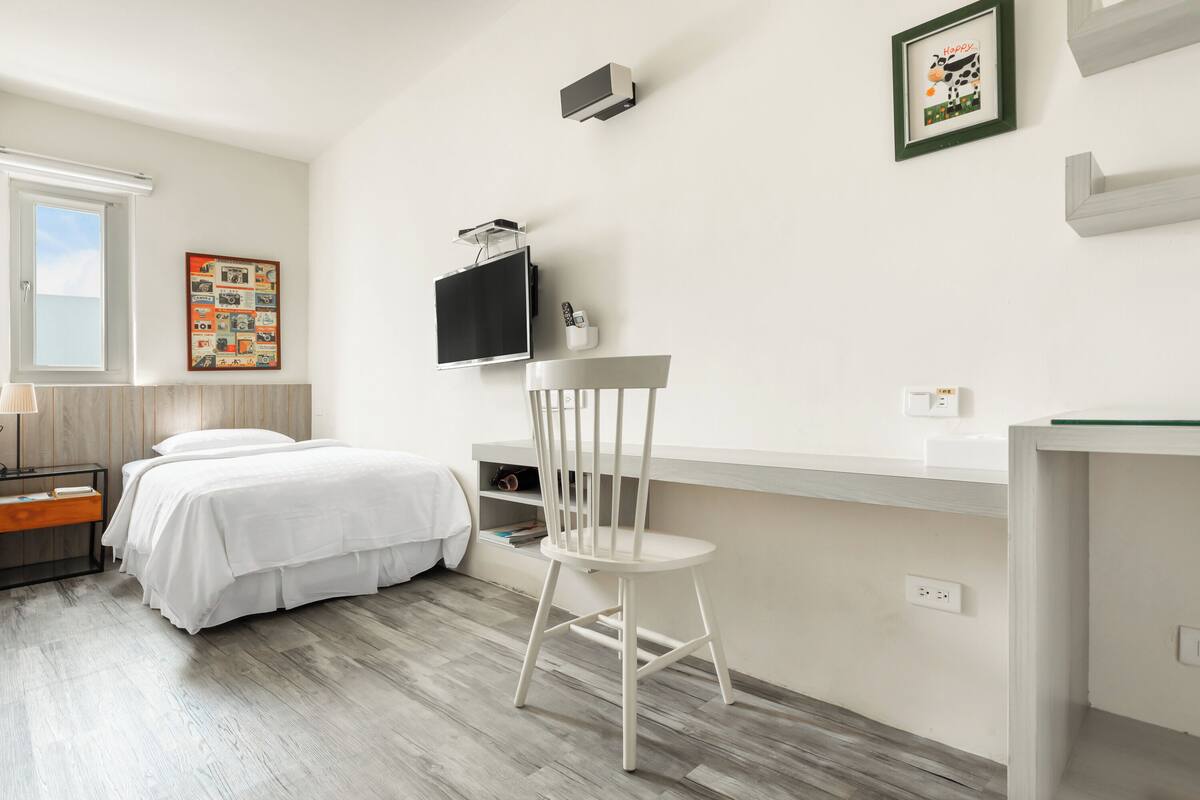 Ein Schlafzimmer mit Bett, Schreibtisch, Stuhl, Fernseher und Fenster.