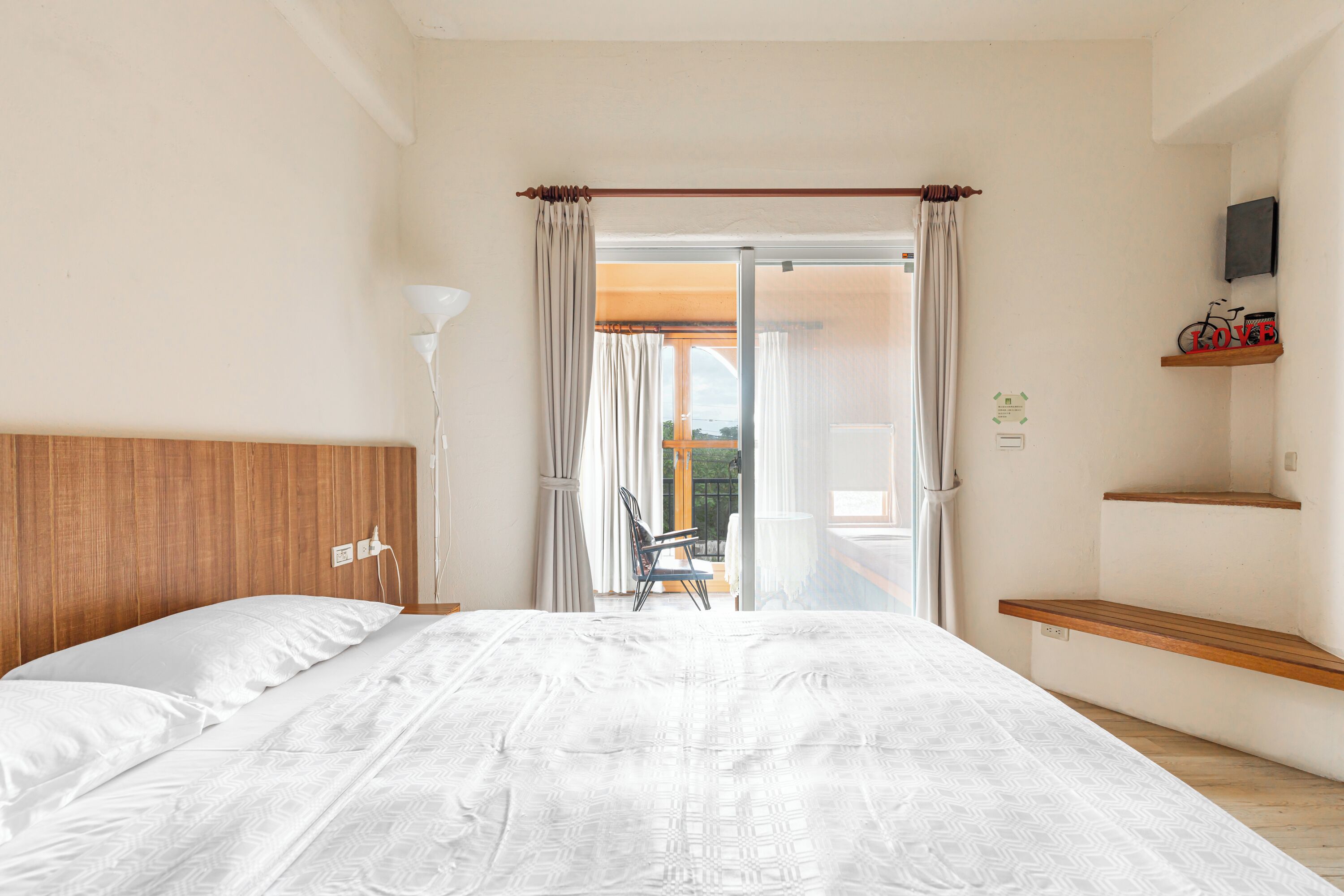 A bedroom with a wooden headboard, a large bed with patterned bedding, a chair, a balcony with a view, and a mounted TV.