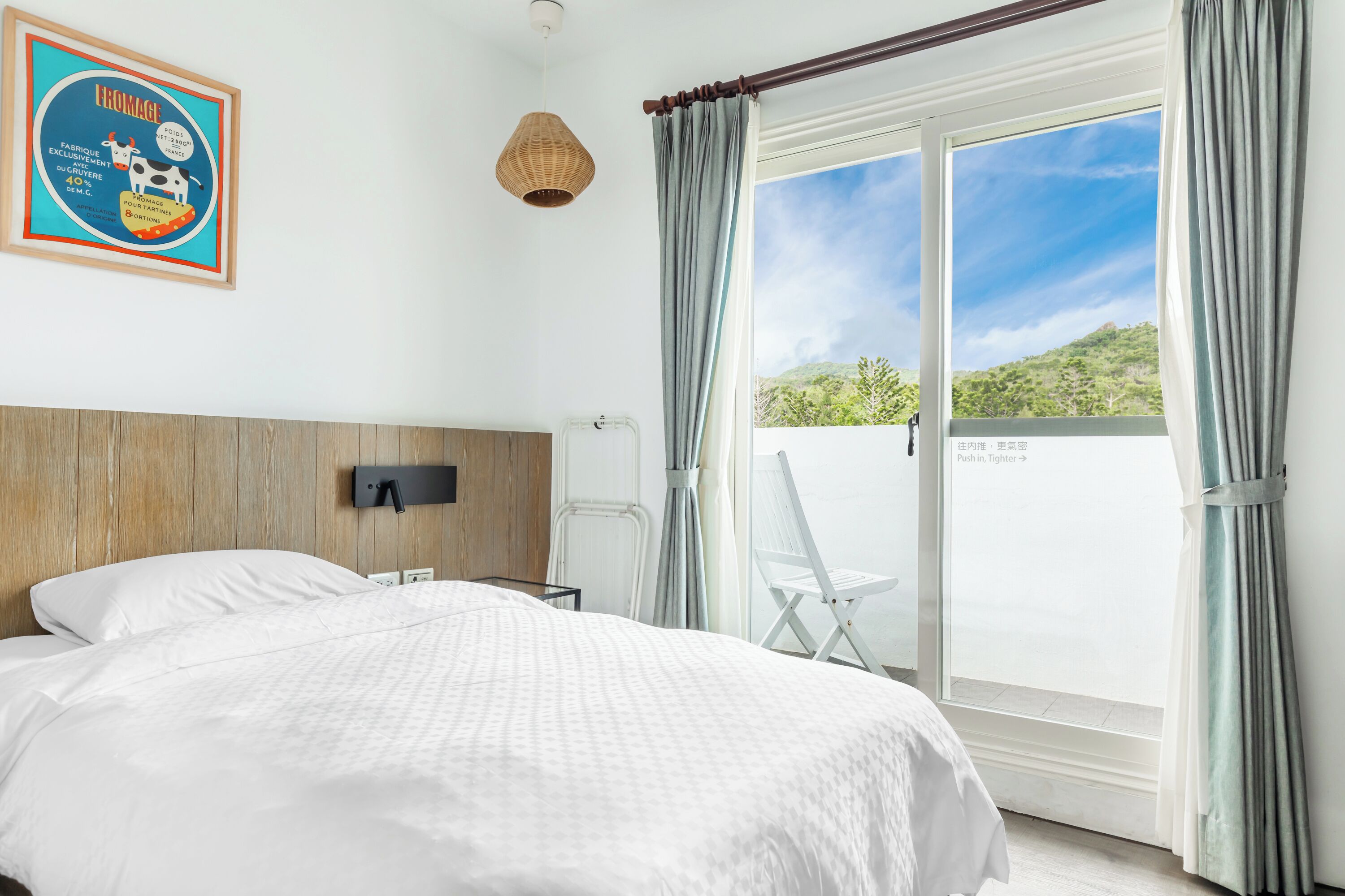 Quarto com uma cama grande, cabeceira de madeira, televisão fixada na parede, luminária de vime, cadeira branca e porta de vidro deslizante que dá para uma varanda com vista para o verde.