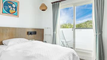 Quarto com uma cama grande, cabeceira de madeira, televisão fixada na parede, luminária de vime, cadeira branca e porta de vidro deslizante que dá para uma varanda com vista para o verde.