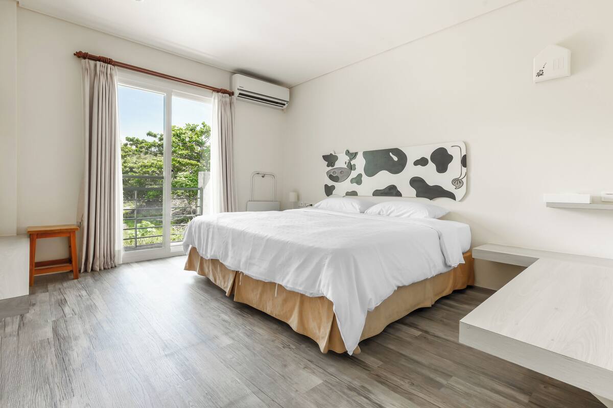 Ein Schlafzimmer mit einem Bett, einem Holznachttisch, einem Beistelltisch, einem Fenster mit Vorhängen und Blick ins Grüne.
