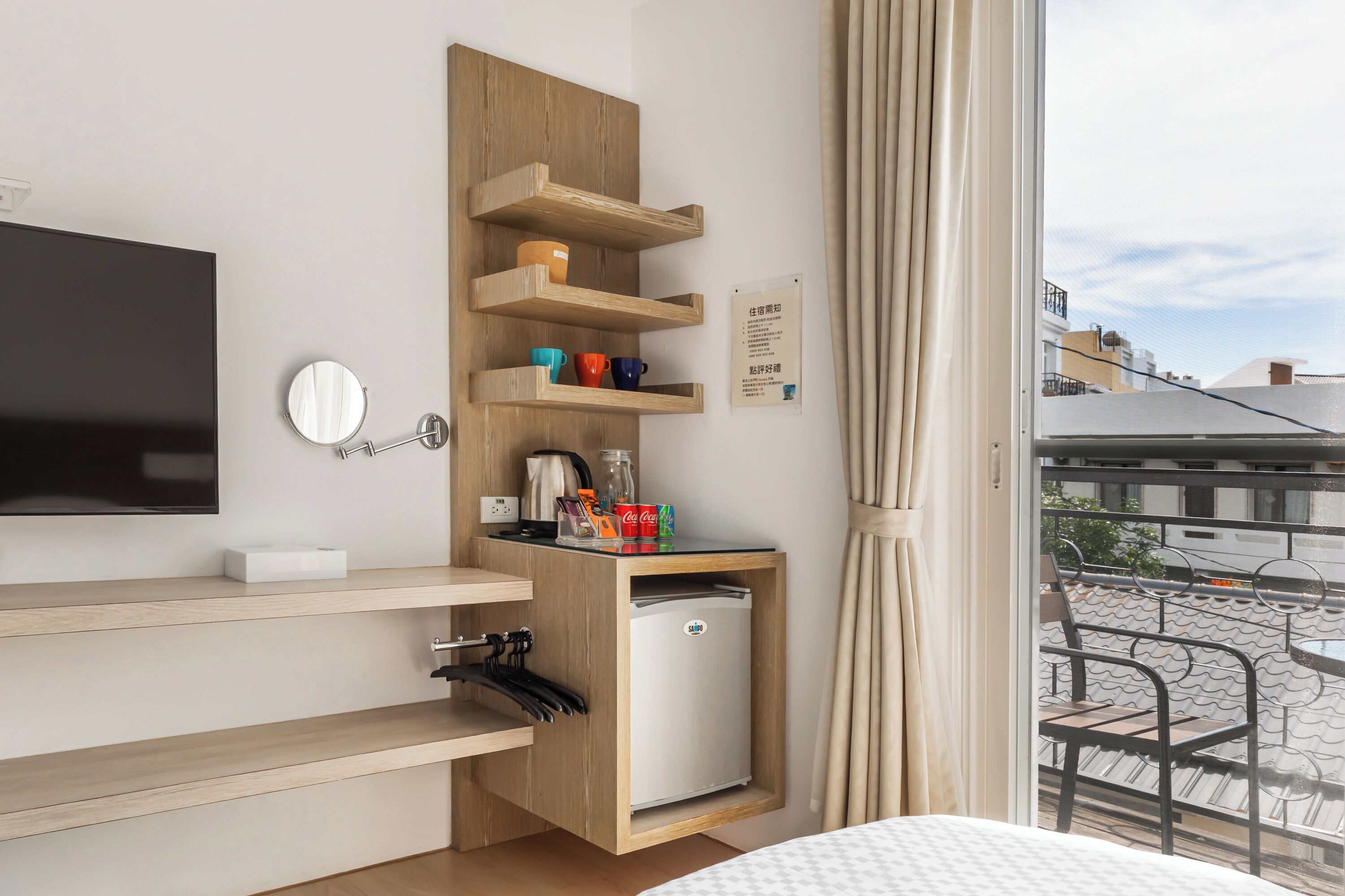 A compact hotel room with a bed, a TV, a small refrigerator, a wooden shelf with cups, and a window with a view of buildings.