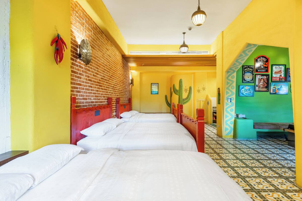 Ein Hotelzimmer mit vier Betten, einer Ziegelwand, einer Kakteen-Dekoration und einer grünen Wand mit verschiedenen Postern.